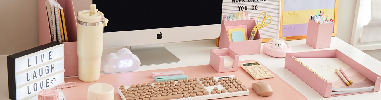 Work or study desk outfitted with neutral-toned desk accessories such as a white keyboard with pink keys, a pink mouse, calculator & desk organizers, markers, pens, gold scissors & more.. 