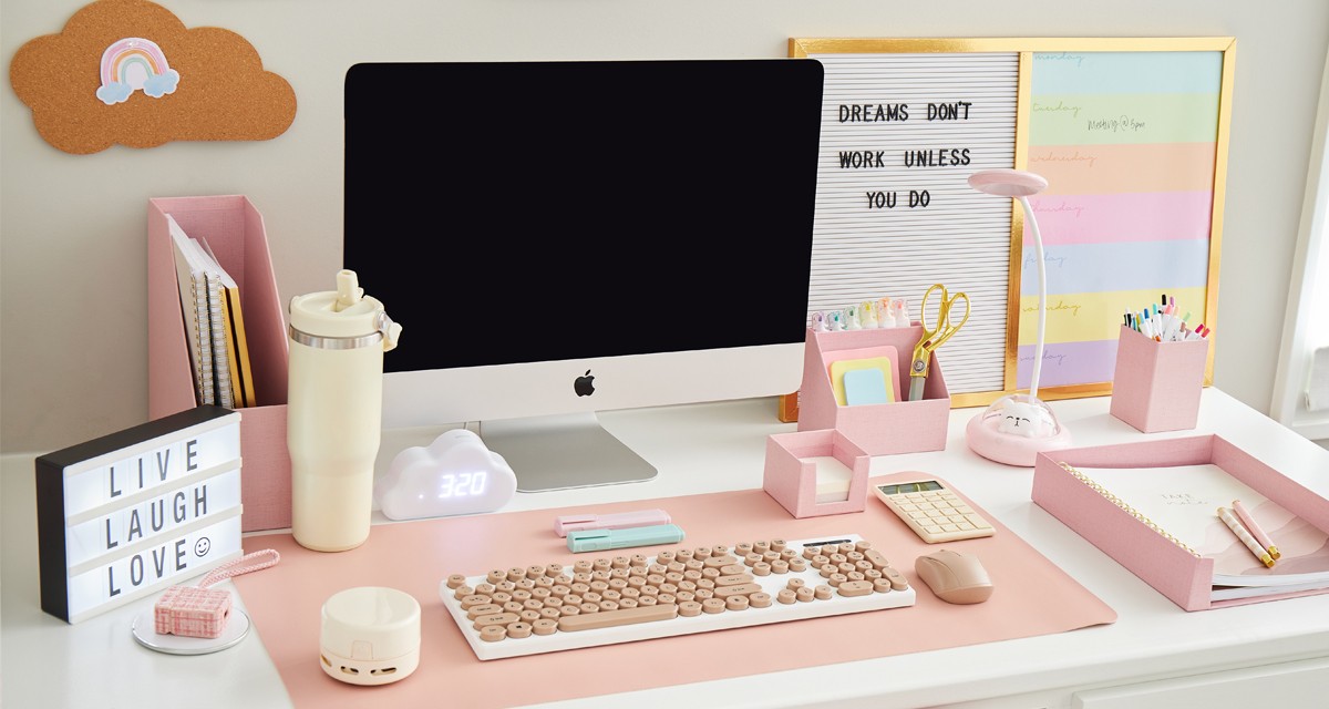 Work or study desk outfitted with neutral-toned desk accessories such as a white keyboard with pink keys, a pink mouse, calculator & desk organizers, markers, pens, gold scissors & more.. 