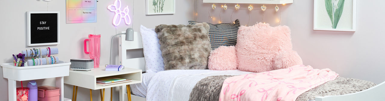 Dorm room decorated with pastel and neautral tones: pink & gray throws, pink furry study pillow, white storage cart, wall lights, sheets & more.
