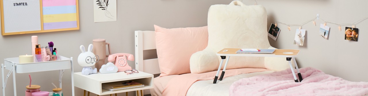 Dorm room decorated with pastel ans neautral tones:pink storage ottoman, pink throw, white study pillow, lap desk, white storage cart, vintage pink phone and more.
