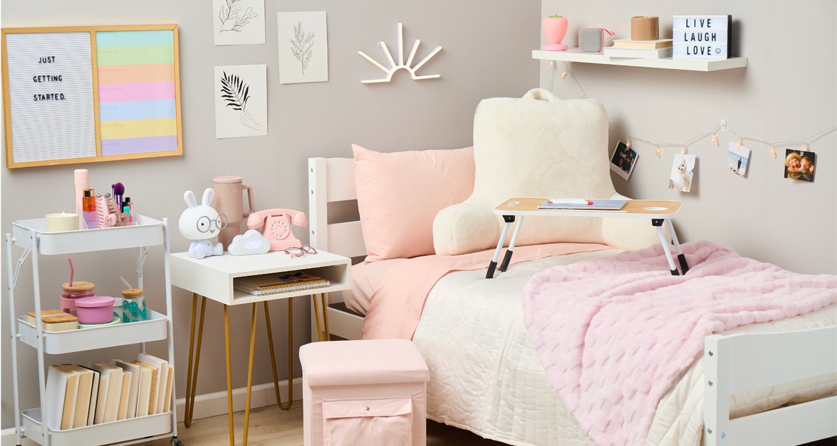 Dorm room decorated with pastel ans neautral tones:pink storage ottoman, pink throw, white study pillow, lap desk, white storage cart, vintage pink phone and more.