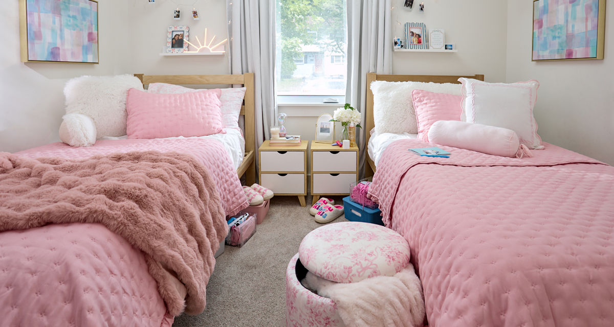 Dorm room decorated with pastel and neautral tones: pink throws, pink furry study pillow, pink floral storage ottomon, wall lights, sheets & more.