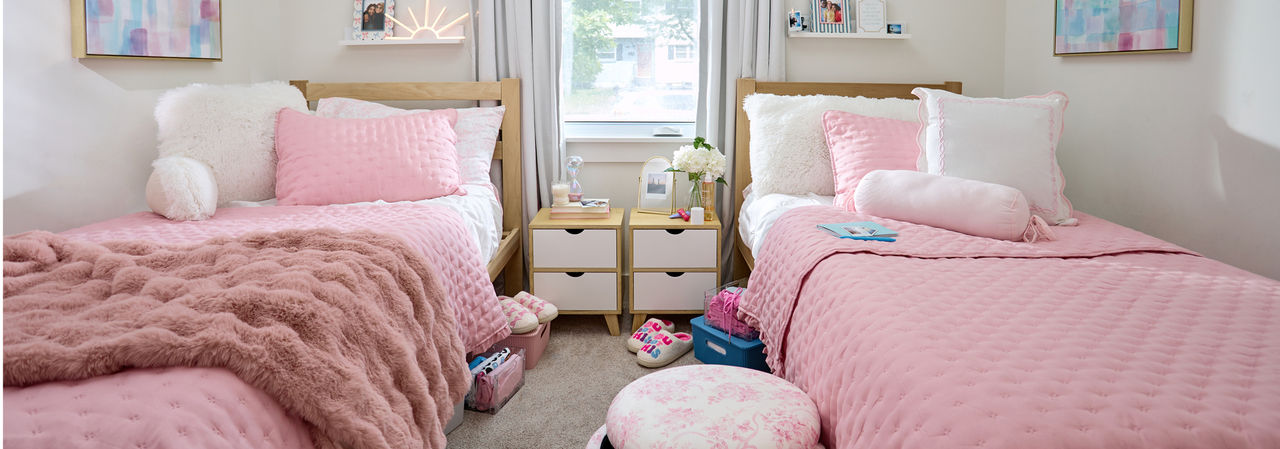 Dorm room decorated with pastel and neautral tones: pink throws, pink furry study pillow, pink floral storage ottomon, wall lights, sheets & more.
