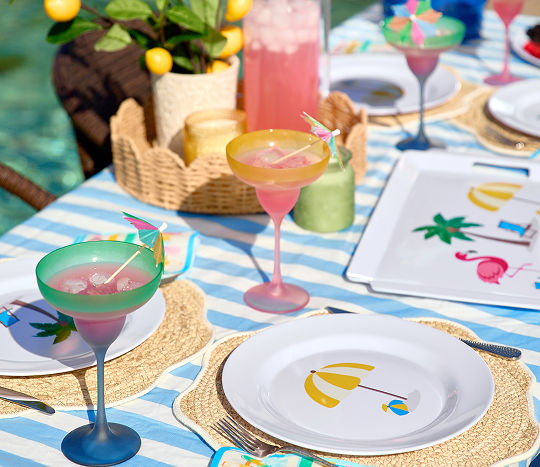 Poolside table set with colorful melamine dinnerware with stripes and summer icons, ice bucket, and matching drinkware.