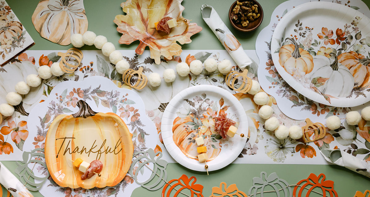Fall party table set with pumpkin and leaf shaped plates, round harvest plates, napkins, gold party utensils, and fall & pumpkin table decor.