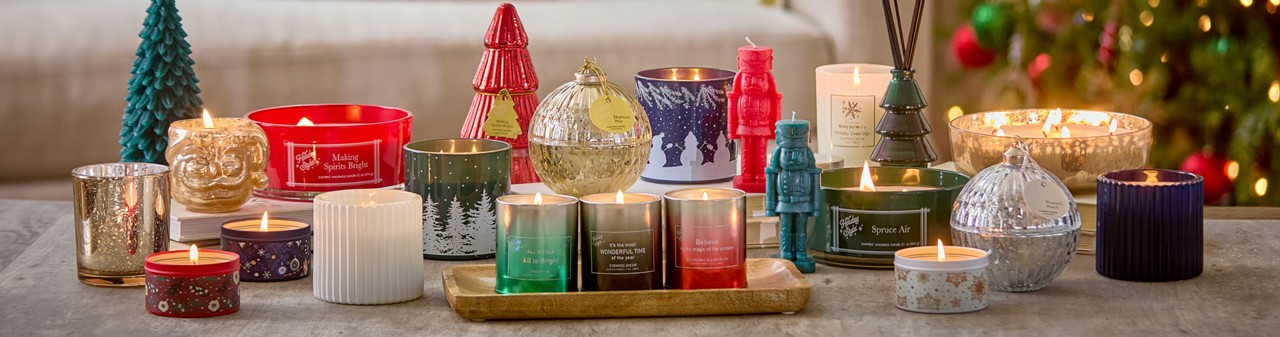Christmas candles in various sizes, designs, and scents on a table in front of a Christmas tree.