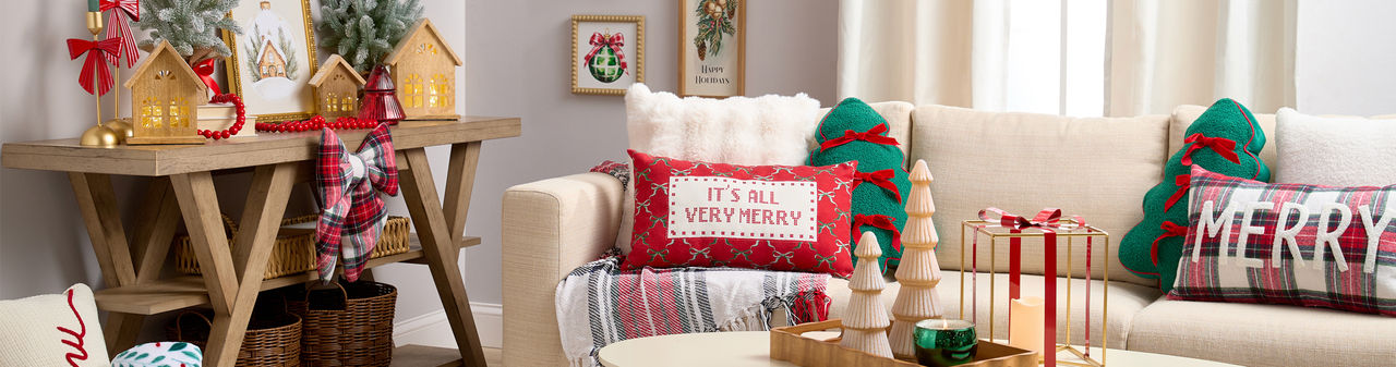 Room decorated for Christmas with wooden trees and village houses, festive pillows & throws, candles, metal lantern, and more. 