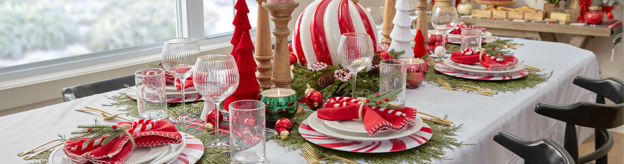Dining table set for Christmas with peppermint candy charger plates and white beaded dinnerware, faux pine placemats, candycane water goblets, and a giant peppermint swirl ornament centerpiece with red and green trees, candles & more.
