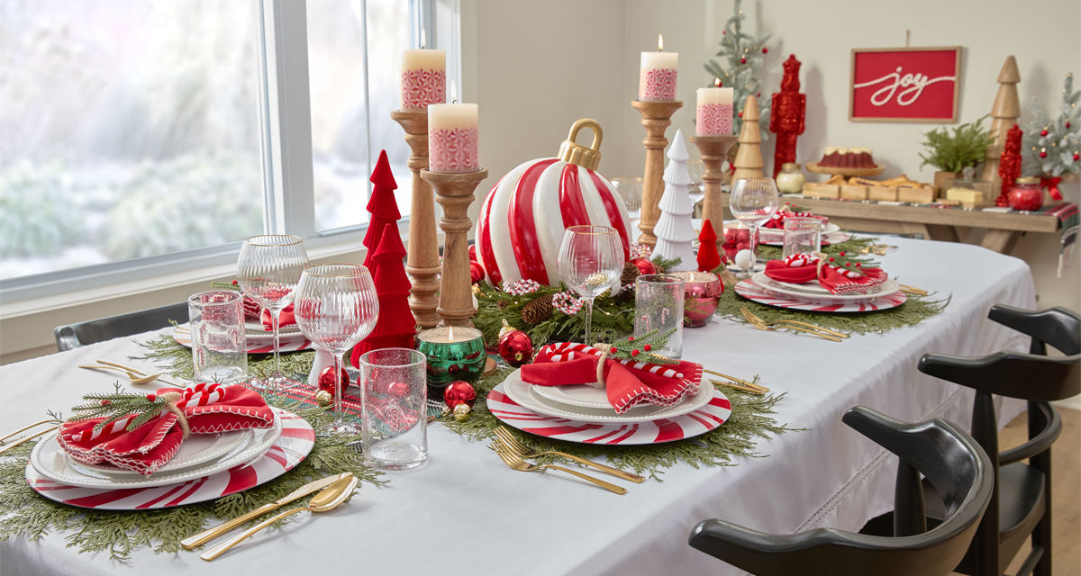 Dining table set for Christmas with peppermint candy charger plates and white beaded dinnerware, faux pine placemats, candycane water goblets, and a giant peppermint swirl ornament centerpiece with red and green trees, candles & more.