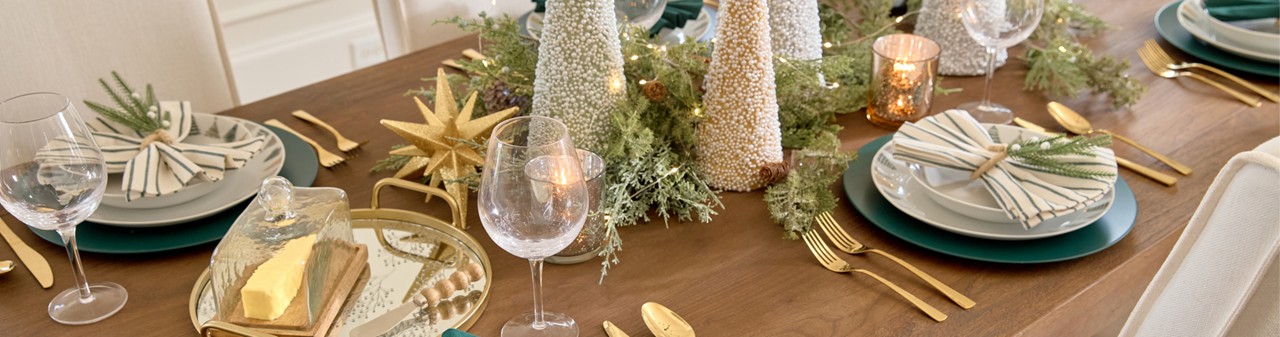 Dining table set for Christmas with ceramic Christmas tree designed dinner plates and bowls, wine glasses, solid and green-striped napkins with pine napkin holders, cone tree centerpiece with faux greenery, lightss, and large sparkly tabletop decor.