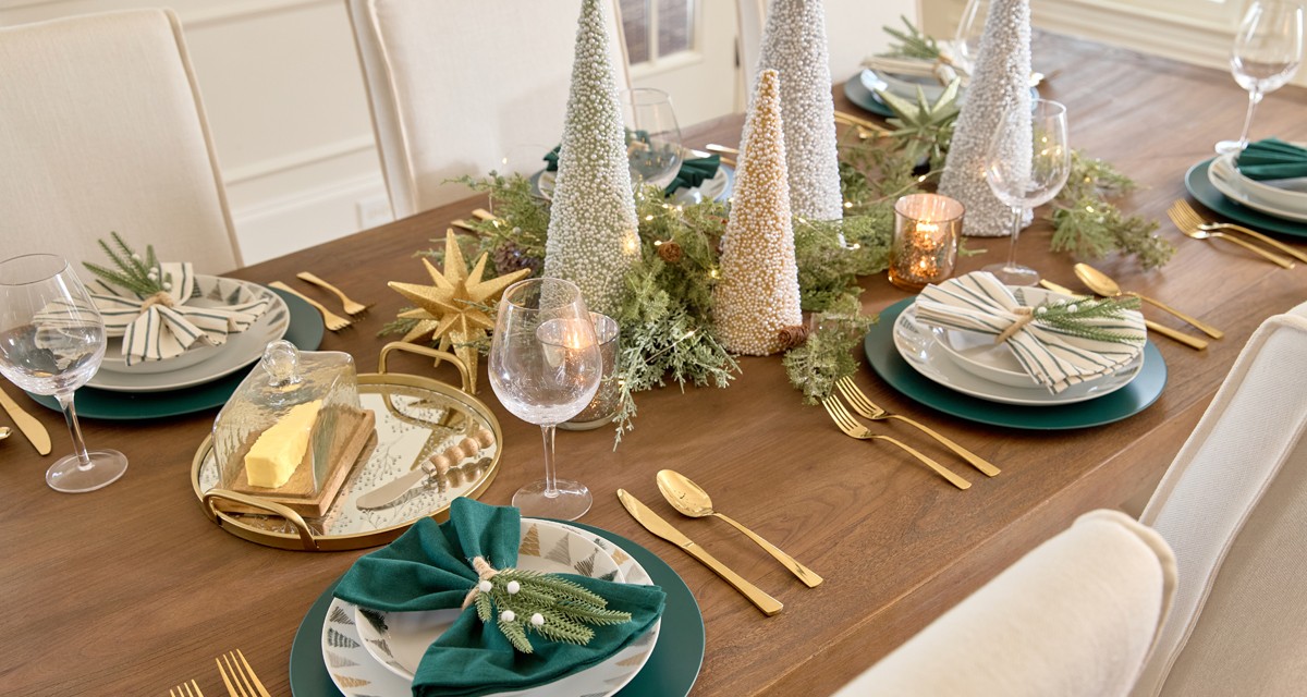 Dining table set for Christmas with ceramic Christmas tree designed dinner plates and bowls, wine glasses, solid and green-striped napkins with pine napkin holders, cone tree centerpiece with faux greenery, lightss, and large sparkly tabletop decor.