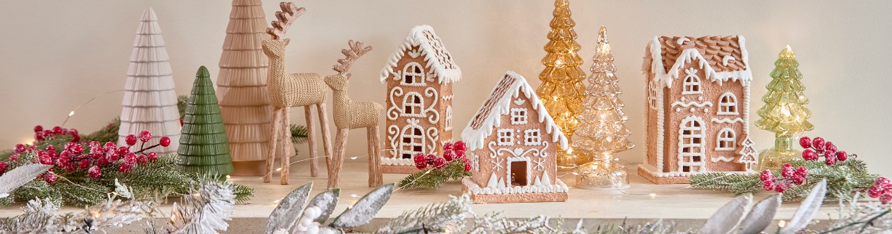 Ceramic gingerbread house decor in three styles on a table with wooden deer and ceranic trees surrounded by Christmas floral, berries & greenery.