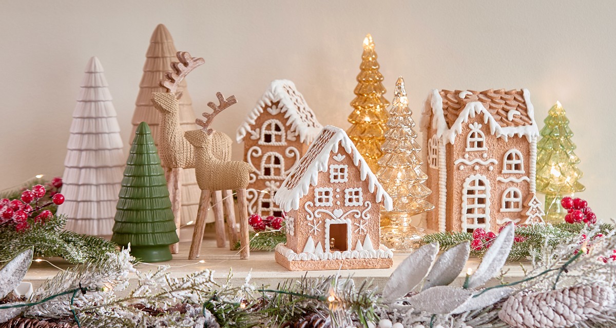 Ceramic gingerbread house decor in three styles on a table with wooden deer and ceranic trees surrounded by Christmas floral, berries & greenery.