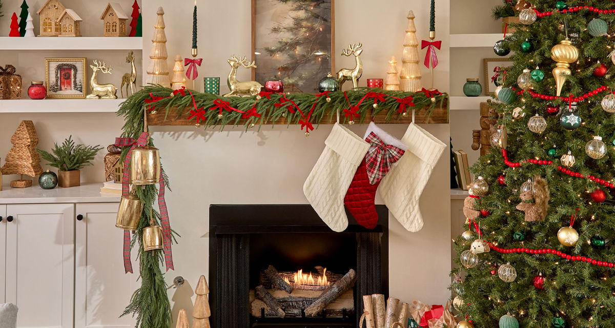 Living room decorated for Christmas with faux pine and large gold bells on thets,  mantel, tree full or ornaments, wooden village houses and trees, stockings, festive Christmas pillows and throws, candles, wall art & more.