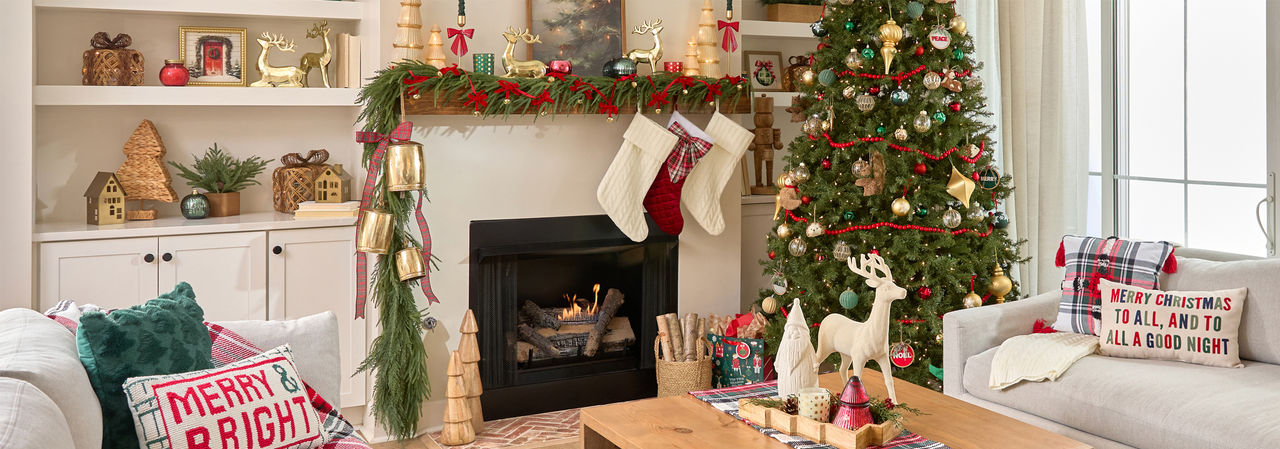 Living room decorated for Christmas with faux pine and large gold bells on thets,  mantel, tree full or ornaments, wooden village houses and trees, stockings, festive Christmas pillows and throws, candles, wall art & more.