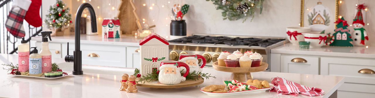 Christmas mixing bowl, wooden cake stand and lazy susan, tree-shaped charcuterie tray, village house cookie jar, Santa mugs, gingerbread man salt & pepper shakers, and more in a kitchen decorated for Christmas.