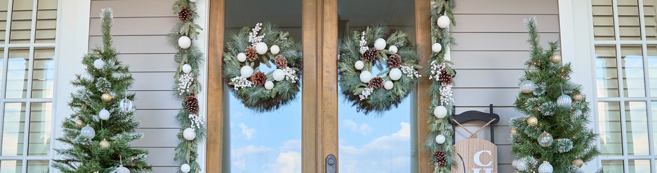 Front porch decorated for Christmas with 6-ft lighted trees, 3-ft. potted trees with fairy lights, sled porcher leaner, wooden trees, lgarland & wreaths, and gold lanterns.