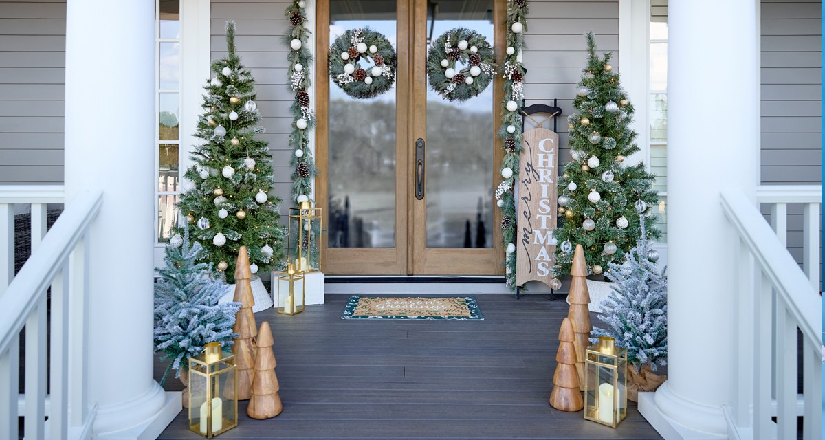 Front porch decorated for Christmas with 6-ft lighted trees, 3-ft. potted trees with fairy lights, sled porcher leaner, wooden trees, lgarland & wreaths, and gold lanterns.