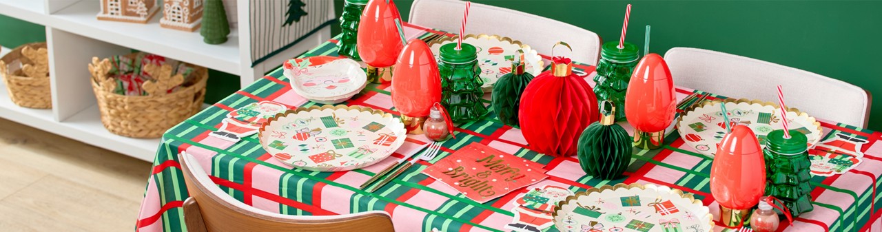 Christmas party table with festive gift and Santa themed party plates and napkins, ornament and glass tree cups with straws, honeycomb ornament centerpiece, and glass ornament drink dispenser.