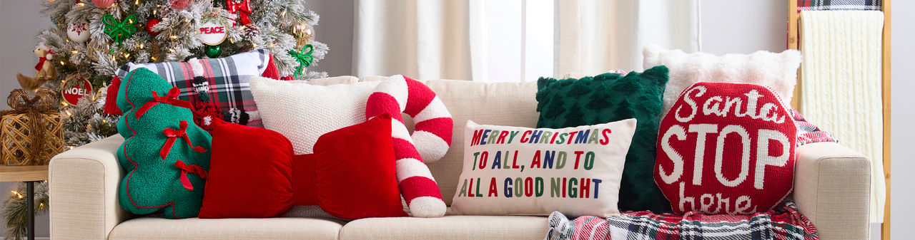 Christmas pillows in various shapes and designs on a sofa: green tree with bows, candy cane, giant red bow, plaid, Santa Stop Here, and more.