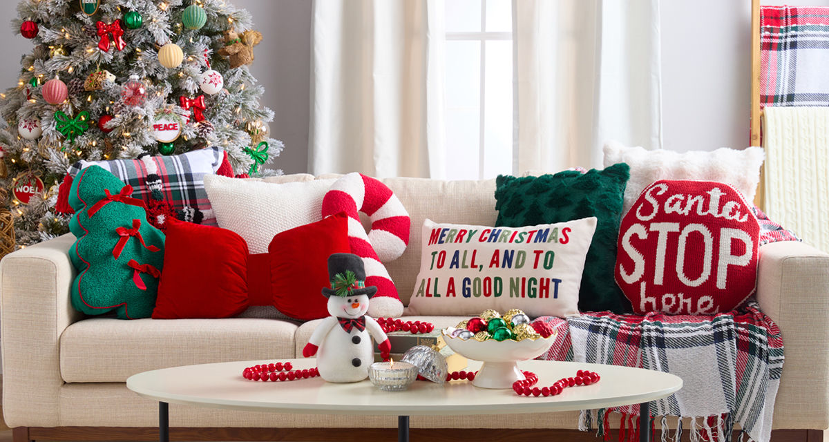 Christmas pillows in various shapes and designs on a sofa: green tree with bows, candy cane, giant red bow, plaid, Santa Stop Here, and more.