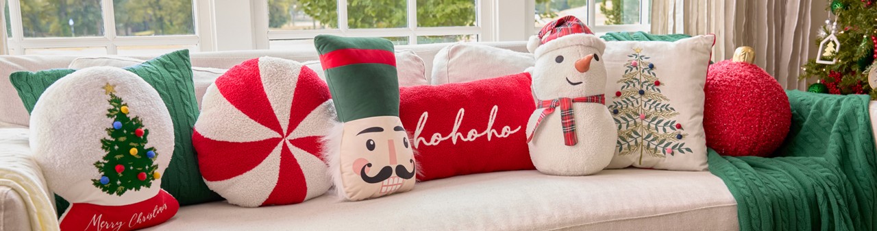 Christmas pillows in various shaped and designs on a sofa: snow globe, peppermint candy, nutcracker, snowman, red ornament & more.
