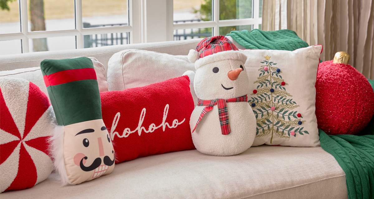 Christmas pillows in various shaped and designs on a sofa: snow globe, peppermint candy, nutcracker, snowman, red ornament & more.