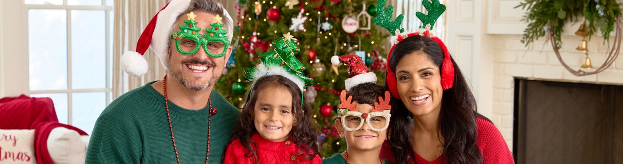 Family in front of Christmas tree wearing Santa hats, novelty Christmas glasses, Christmas bead necklaces, antler and tree headbands, and more.