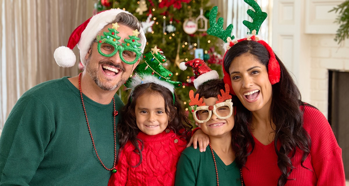 Family in front of Christmas tree wearing Santa hats, novelty Christmas glasses, Christmas bead necklaces, antler and tree headbands, and more.