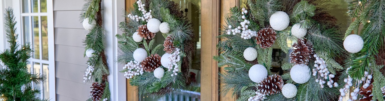 Front door with two faux floral wreaths and garland featuring white ornaments, flocked pine and pinecones, and white berries.