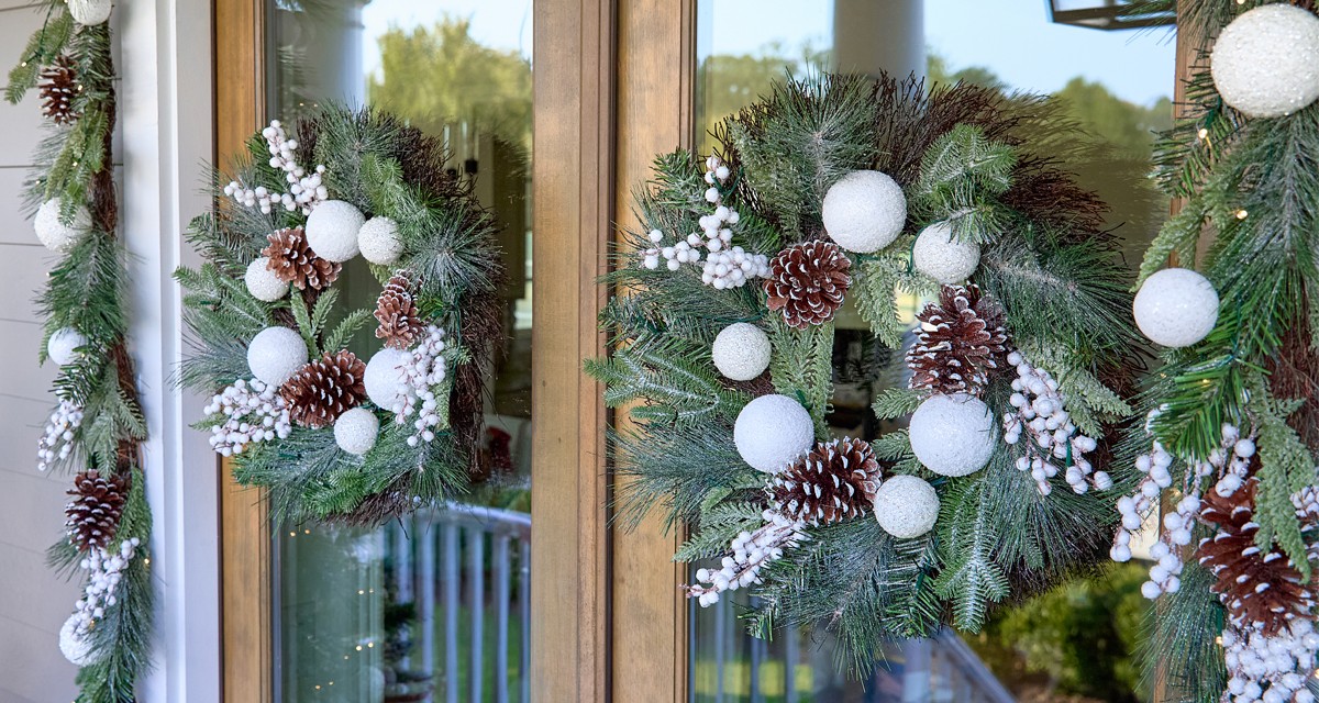 Front door with two faux floral wreaths and garland featuring white ornaments, flocked pine and pinecones, and white berries.