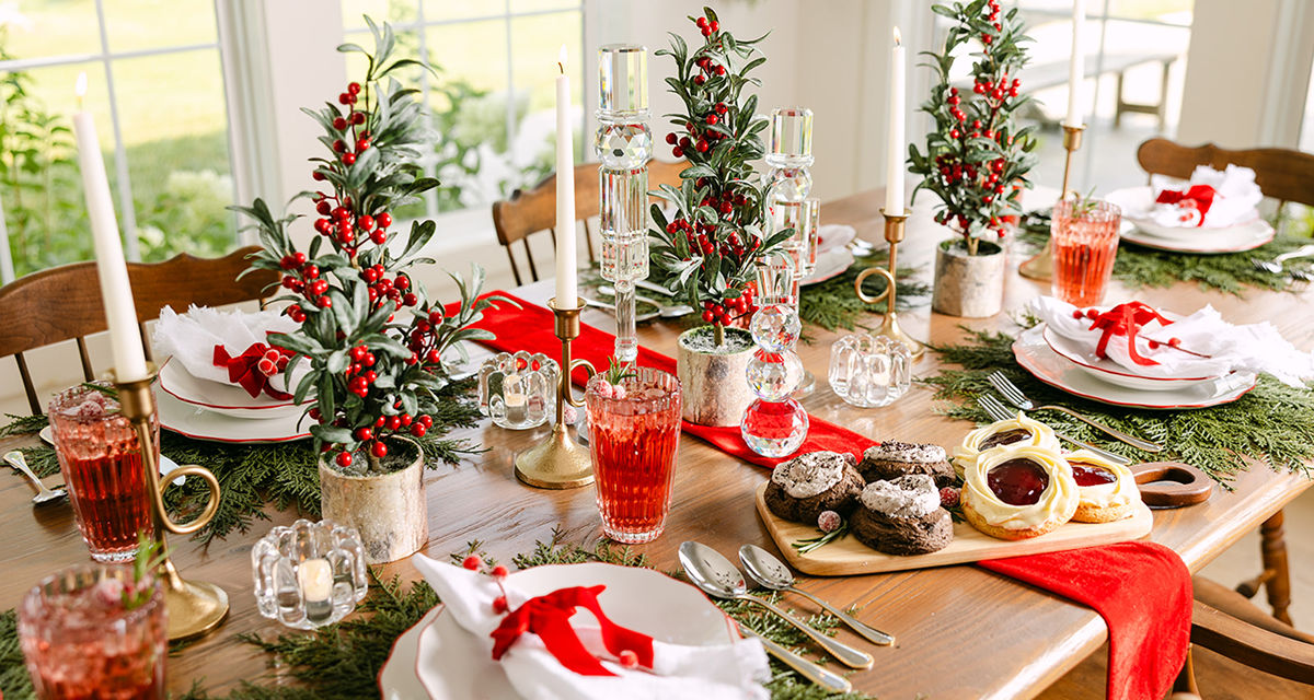 Dining table set for Christmas with faux pine chargers, red trimmed dinnerware, water goblets, and a giant peppermint swirl ornament centerpiece with red and green trees, candles & more.