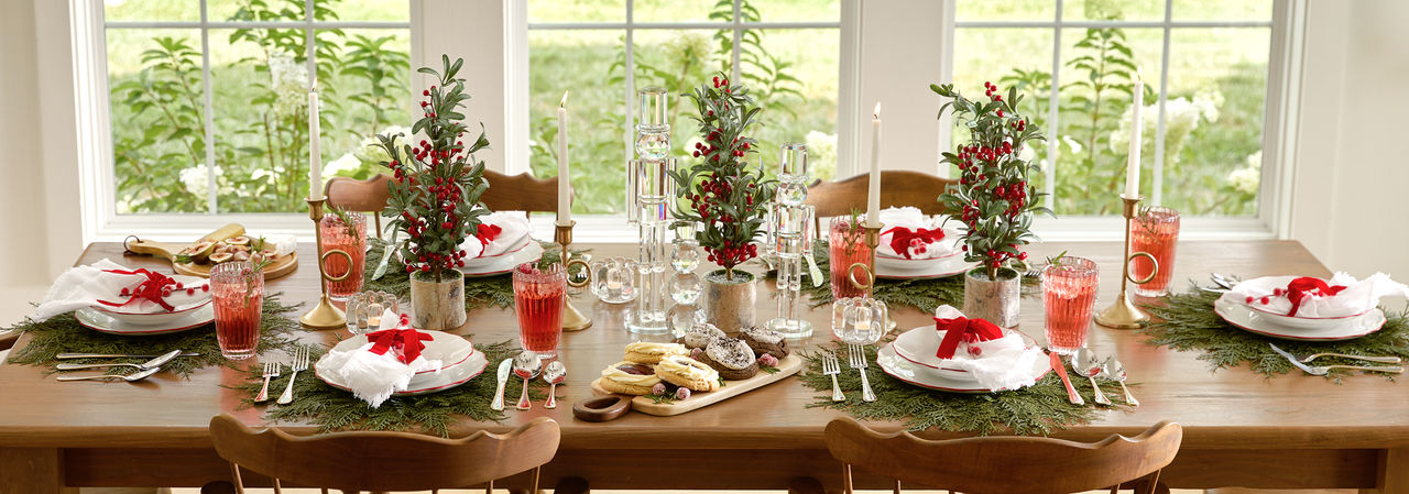 Dining table set for Christmas with faux pine chargers, red trimmed dinnerware, water goblets, and a giant peppermint swirl ornament centerpiece with red and green trees, candles & more.