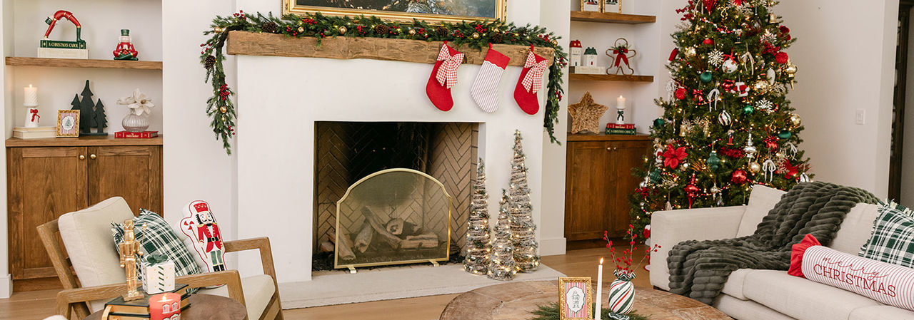 Living room decorated for Christmas with faux pine garland, stockings, tree full or ornaments, festive Christmas pillows and throws, candles, tabletop decor & more.