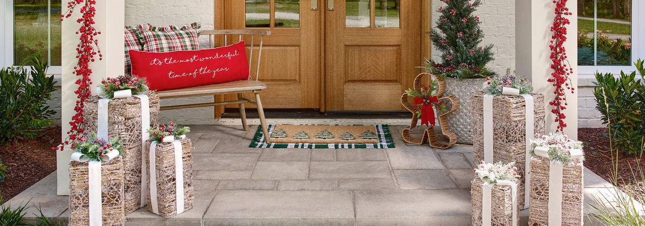 Front porch decorated for Christmas with wreaths garland, coir mats, rattan decor, pillows, bells & more.