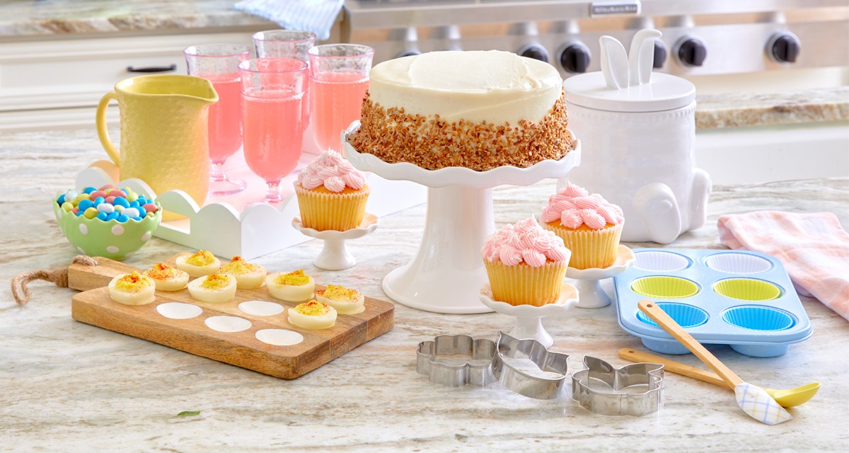 Easter kitchen with wooden egg tray, yellow ceramic pitcher, white cake stand, white bunny-ear canisters, glass goblets, mini dessert stands, Easter decor, baking items & more.