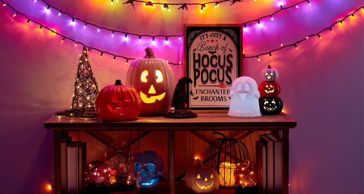 Halloewen string lights and light-up witch's hat, wicker pumpkin, jack-o-lanterns, ghosts & more on an entryway table.
