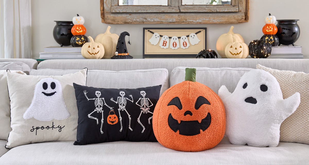 Halloeen pillows lined up on a sofa: square ghost illow, black pillow with skeletons, and ghost- & Jack-o-lantern-shaped pillows.