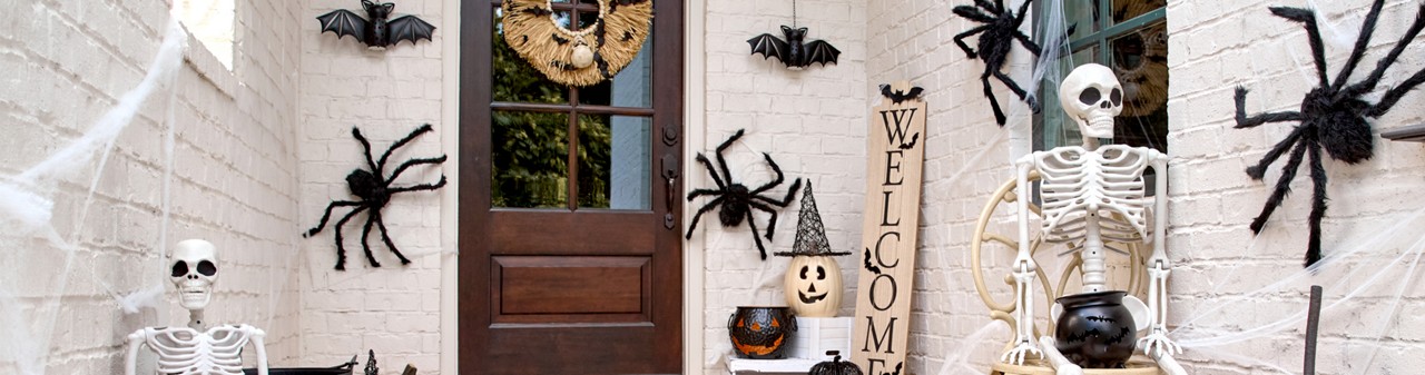 Front porch decorated for Halloween with giant spiders, skeletons, pumpkins and jack-o-lanterns, solar bat lanterns, witches hats, and a Welcome porch leaner.