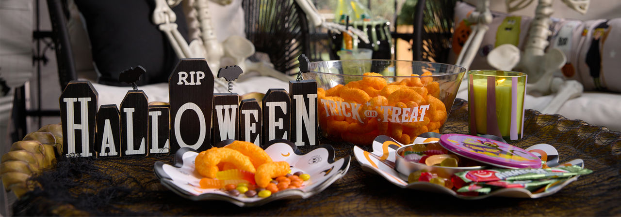A porch is set with two faux skeletons sitting in lounge chairs with a table decorated with Halloween candy and snacks and decorated with fall florals and a classic Halloween Jack-O-Lantern themed pumpkin.