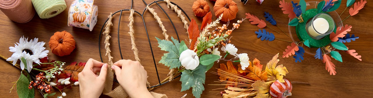 Woman completing wire pumpkin wreath form with fall florals & picks, fall ribbon, a DIY fall centerpiece, and pumpkin embellishments.