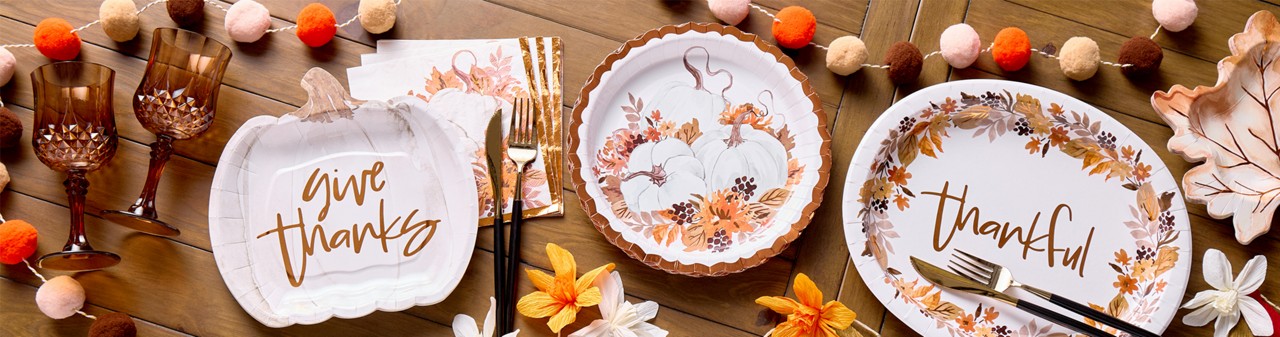 Fall party supplies: pom-pom garland, pumpkin- and leaf-shaped plates, plates and napkins with a pumpkin design, plastic brown wine glasses & more.