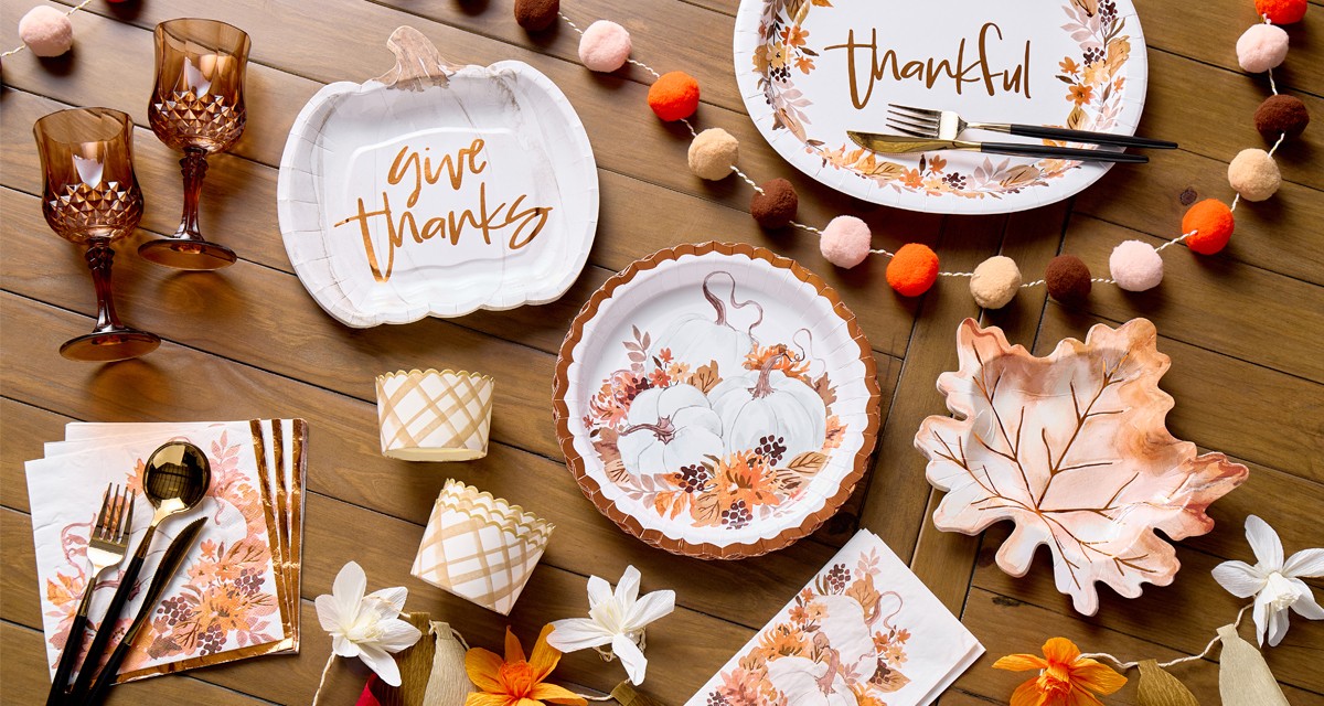 Fall party supplies: pom-pom garland, pumpkin- and leaf-shaped plates, plates and napkins with a pumpkin design, plastic brown wine glasses & more.