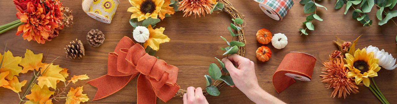 Woman completing wire pumpkin wreath form with fall florals & picks, fall ribbon, a DIY fall centerpiece, and pumpkin embellishments.