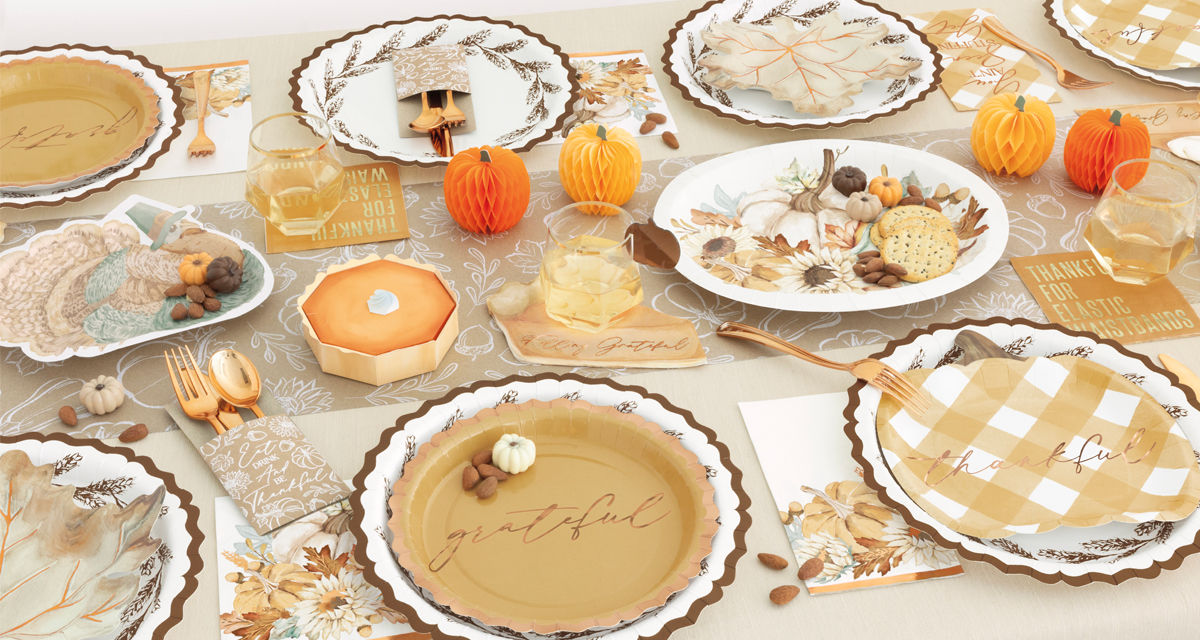 Fall party table set with pumpkin, turkey, and leaf shaped plates, round harvest plates, napkins, gold party utensils, and fall & pumpkin table decor.