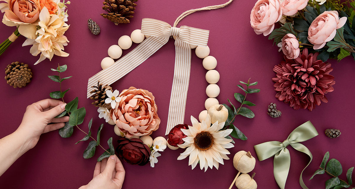 Woman decorating a bead wreath with fall florals & picks, fall ribbon, and pumpkin embellishments.