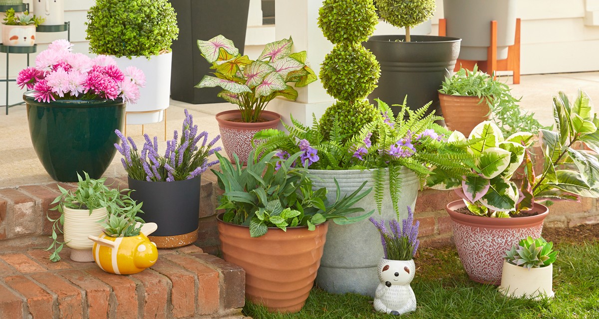 Various colors and designs of pOpshelf outdoor planters, flower pots, and plant stands filled with artificial greenery and flowers on a front porch.