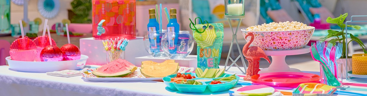 Poolside table set with colorful Palm Springs Pool Party supplies: drink dispenser, paper plates and napkins, fish bowl cocktail glasses with mixers, melamine tray and serving bowl, and more.