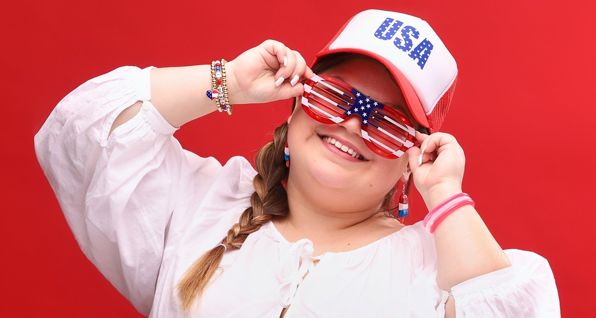 Woman wearing red, white & blue patrioc apparel & accessories from pOpshelf: USA trucker hat, patriotic stretchy bracelets, popsicle earrings, and light-up flag glasses.