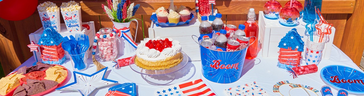 Table set with red, white & blue patriotic party supplies from pOpshelf: star paper plates, cups, napkins, utensils, rocket pinatas, popcorn holders, metal drink bucket, disco ball cups, and more.